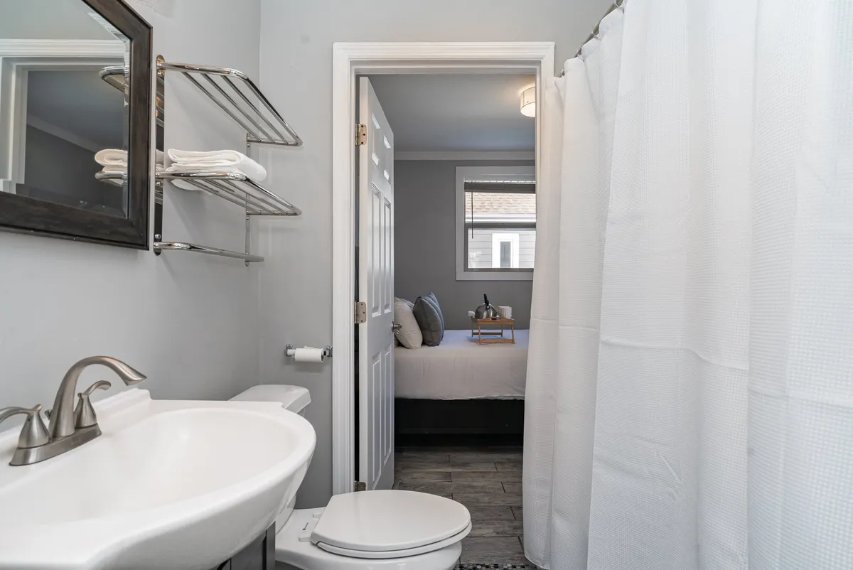 A bright and organized bathroom scene featuring a sink, large mirror, shower curtain, and a doorway leading into a bedroom, showcasing a functional and calming space.