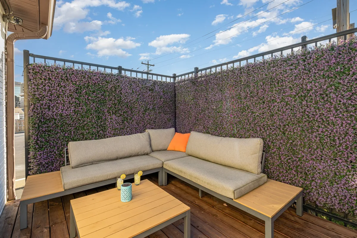 A cozy, fenced outdoor nook featuring a sectional couch with a colorful cushion, a wooden coffee table, and a decorative plant wall. The space invites both relaxation and socializing, with greenery providing extra privacy and serenity—ideal for coffee mornings or evening conversations under the stars.