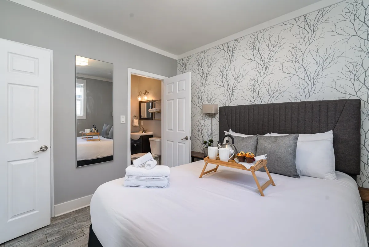 A cozy bedroom with a bed adorned with decorative pillows, a breakfast tray for morning indulgence, a mirrored wall, and patterned wallpaper, exuding warmth and inviting calm.