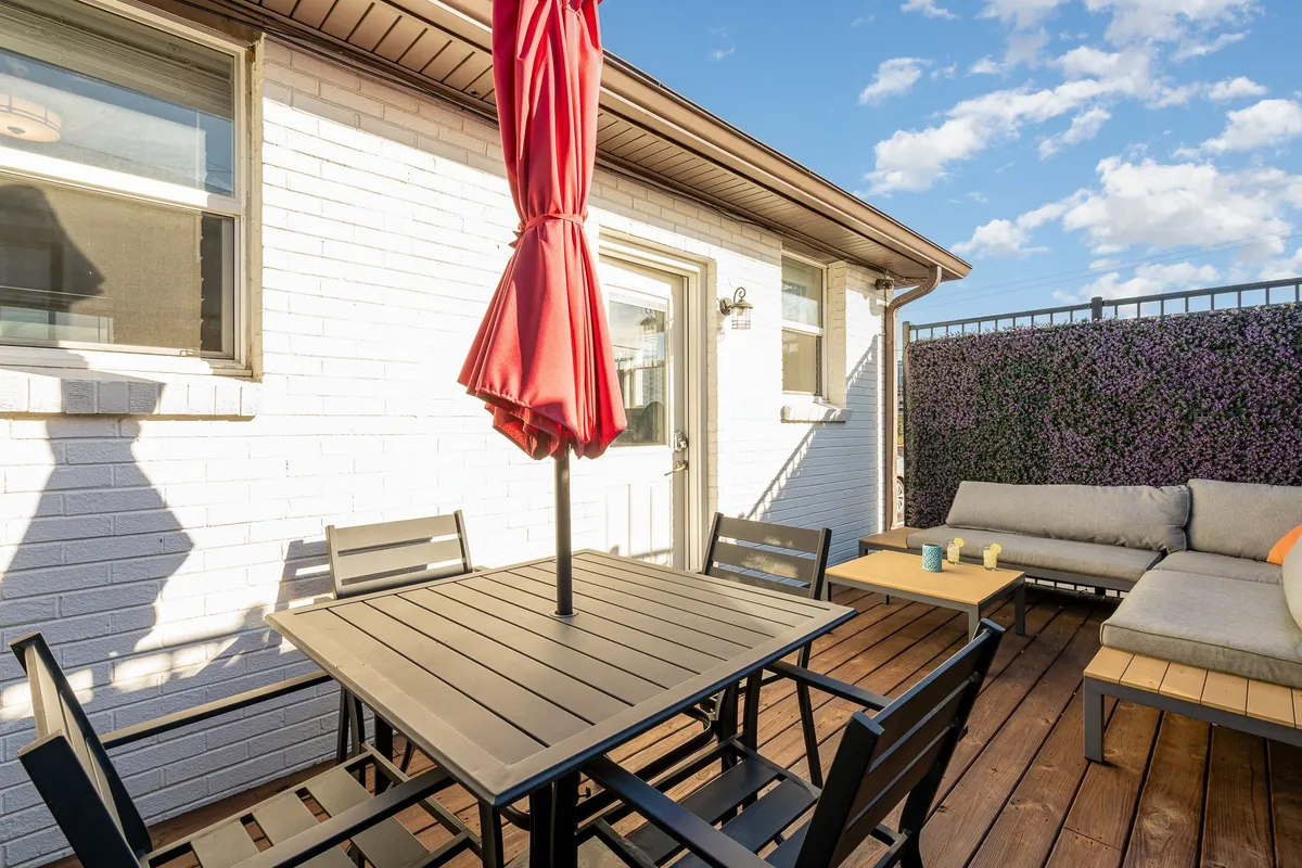 The photo features a contemporary patio area with a stylish red umbrella, a modern outdoor dining table for four, and inviting seating. The setting highlights space for alfresco meals and casual gatherings, with a decorative wall adding a touch of character to the outdoor oasis.