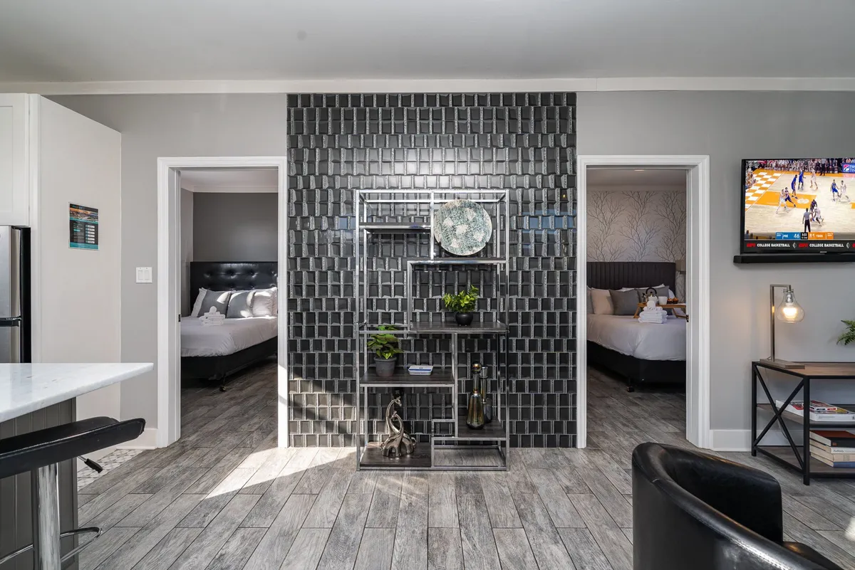 A sleek living area with a black tiled wall, open doorways leading to adjacent rooms, a stylish shelving unit, and a television. The space is arranged for both relaxation and entertaining guests, emphasizing a modern, cozy atmosphere.